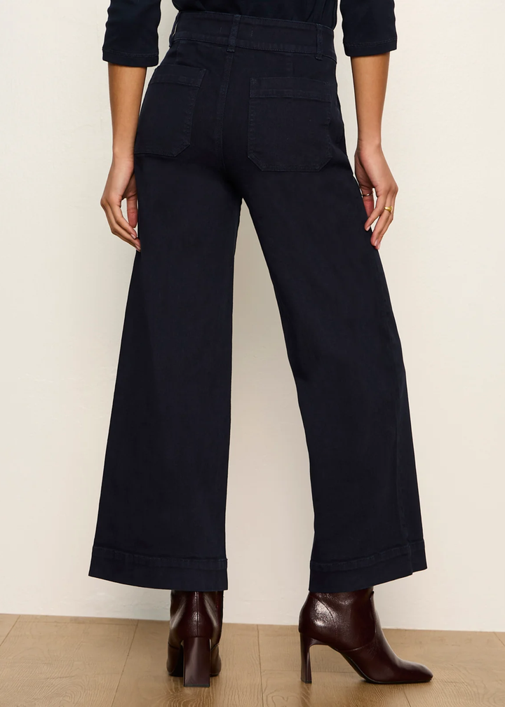A person shown from the back wearing dark navy wide-leg cropped pants with a cuffed hem, paired with dark brown ankle boots against a plain light beige wall and wood floor.