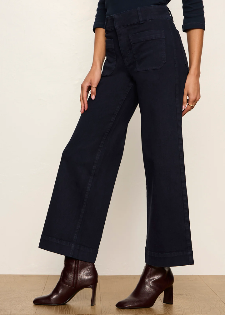A model wearing dark navy wide-leg cropped jeans with a high waist and front patch pockets, paired with dark brown heeled ankle boots against a plain beige studio background.