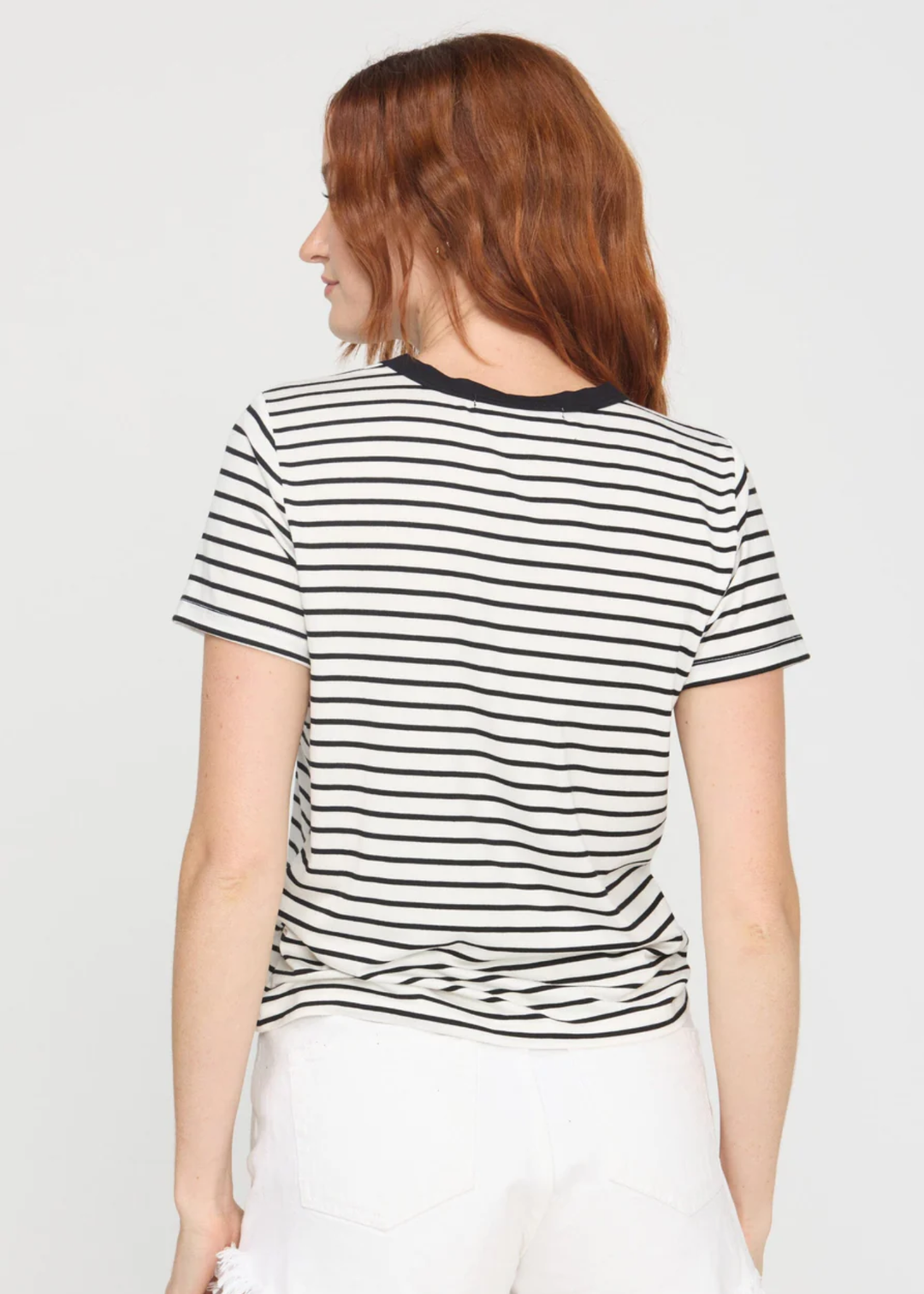 A woman with red hair is shown from the back wearing a short-sleeve black and white horizontally striped t-shirt with a black crew neck, paired with white pants, against a light gray background.
