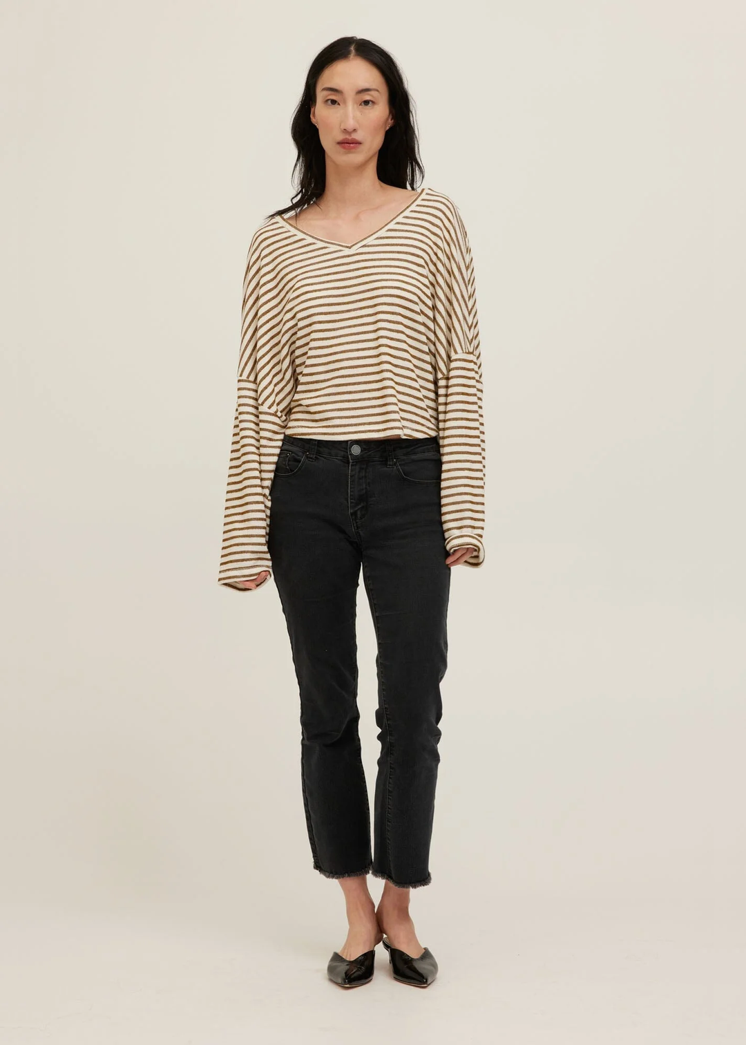 A woman modeling a beige and black horizontally striped long-sleeve V-neck top with black cropped jeans and black pointed flats, standing against a plain light beige studio background.
