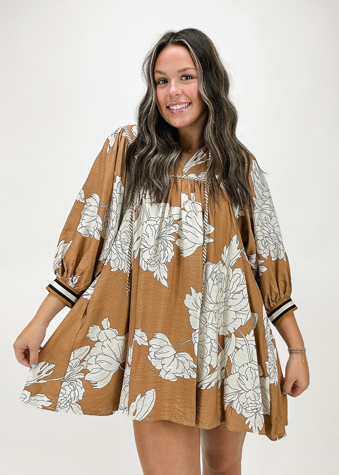 A woman wearing a tan mini dress with large white floral prints, featuring billowy 3/4 sleeves and a drawstring neckline, perfect for a flowy, effortless style.