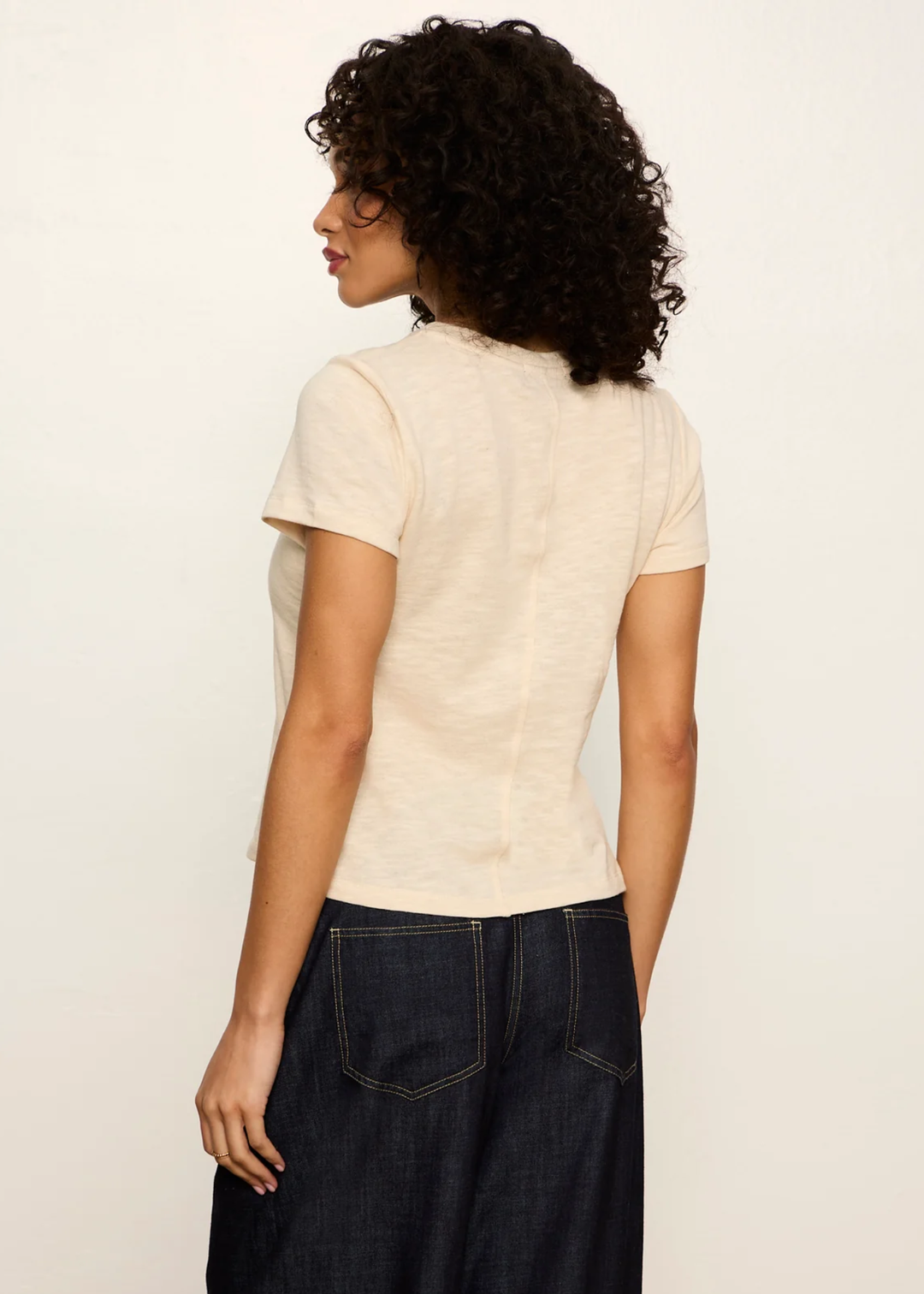A woman with curly dark hair is shown from the back wearing a short-sleeve cream-colored top and dark denim jeans against a plain off-white background.