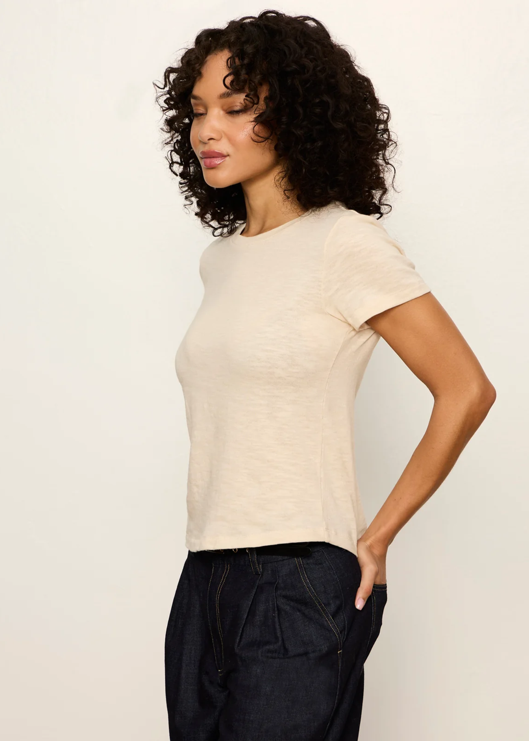 A woman with curly dark hair wearing a light beige short-sleeve crewneck t-shirt and dark denim pants, posing against a plain off-white background.