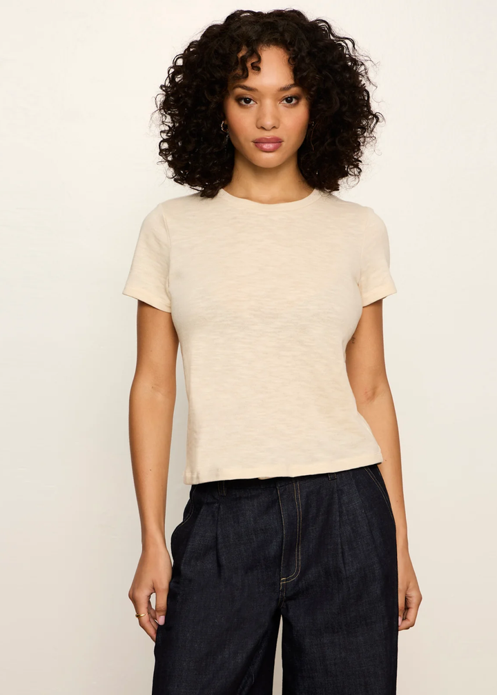 A woman with curly black hair wearing a short-sleeve crewneck t-shirt in a light cream color and dark wide-leg jeans, standing against a plain off-white background.