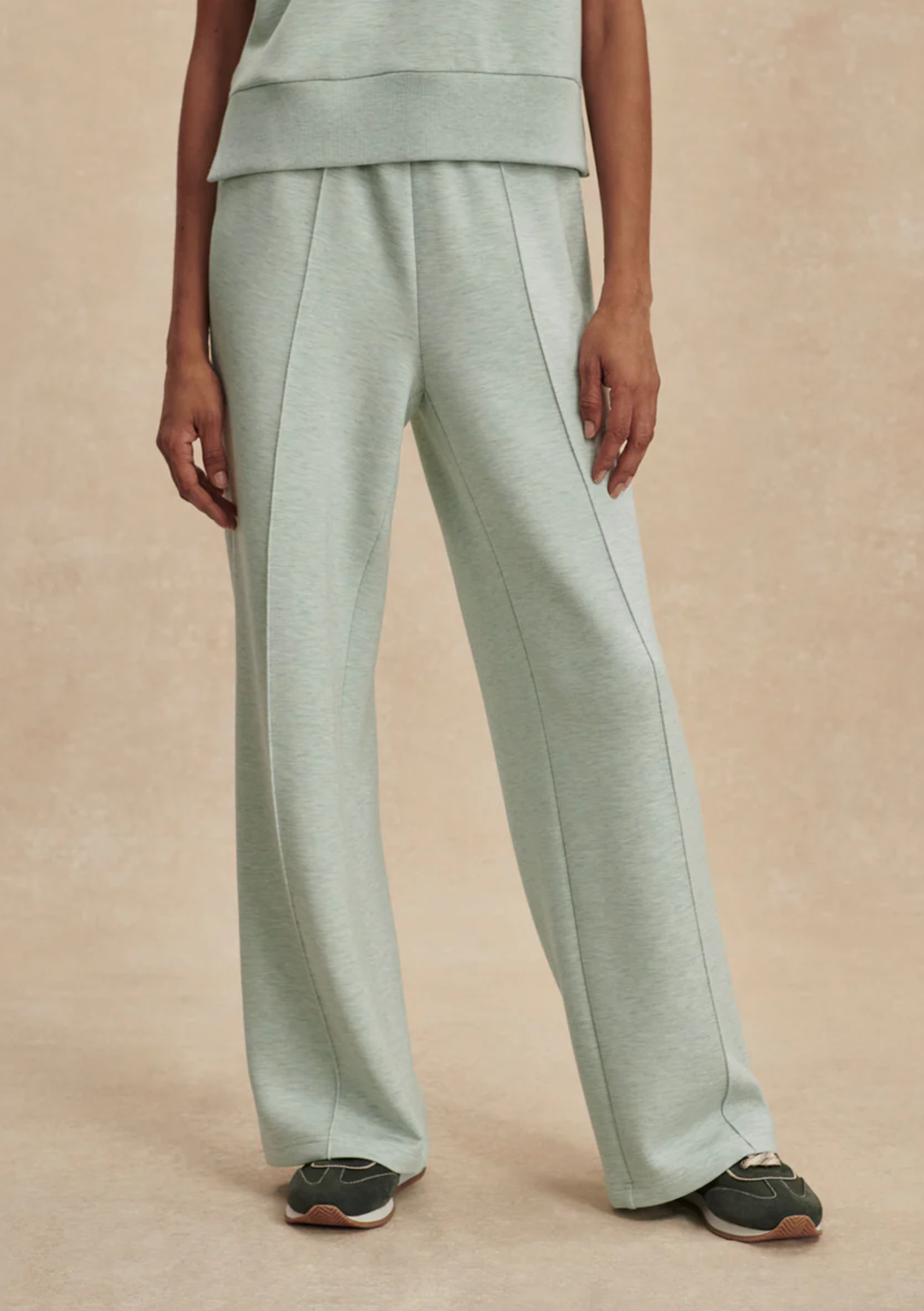 A person wearing light sage green wide-leg sweatpants with a matching cropped top, shown from the waist down against a warm beige studio background.