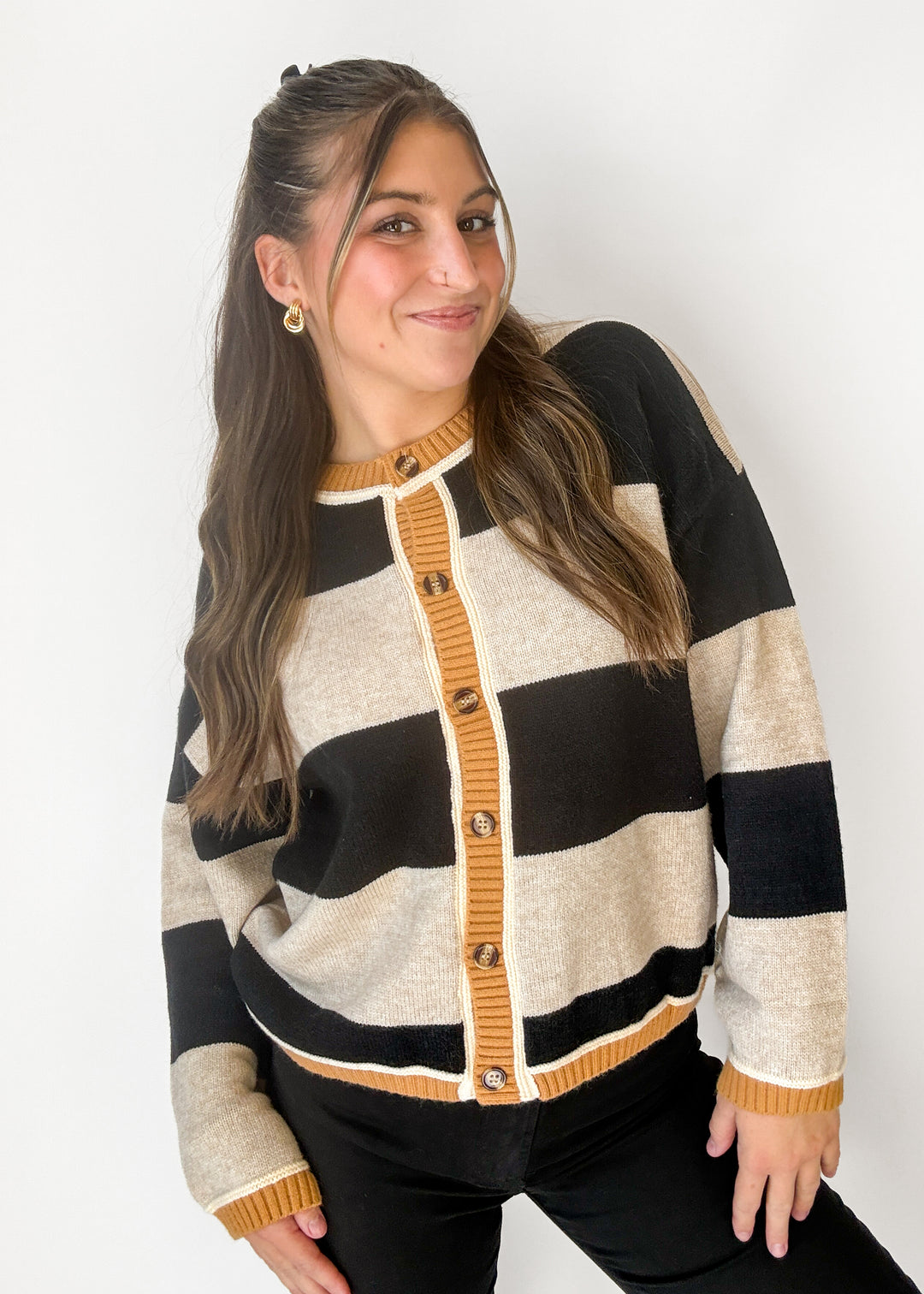 A woman wearing a knit cardigan sweater with wide horizontal stripes in black, cream, and mustard yellow, featuring a mustard yellow button placket and ribbed trim.