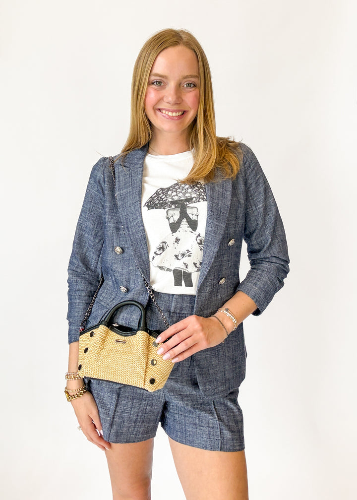 Woman wearing a blue checkered blazer and shorts with a white graphic t-shirt, holding a straw handbag on a white background.