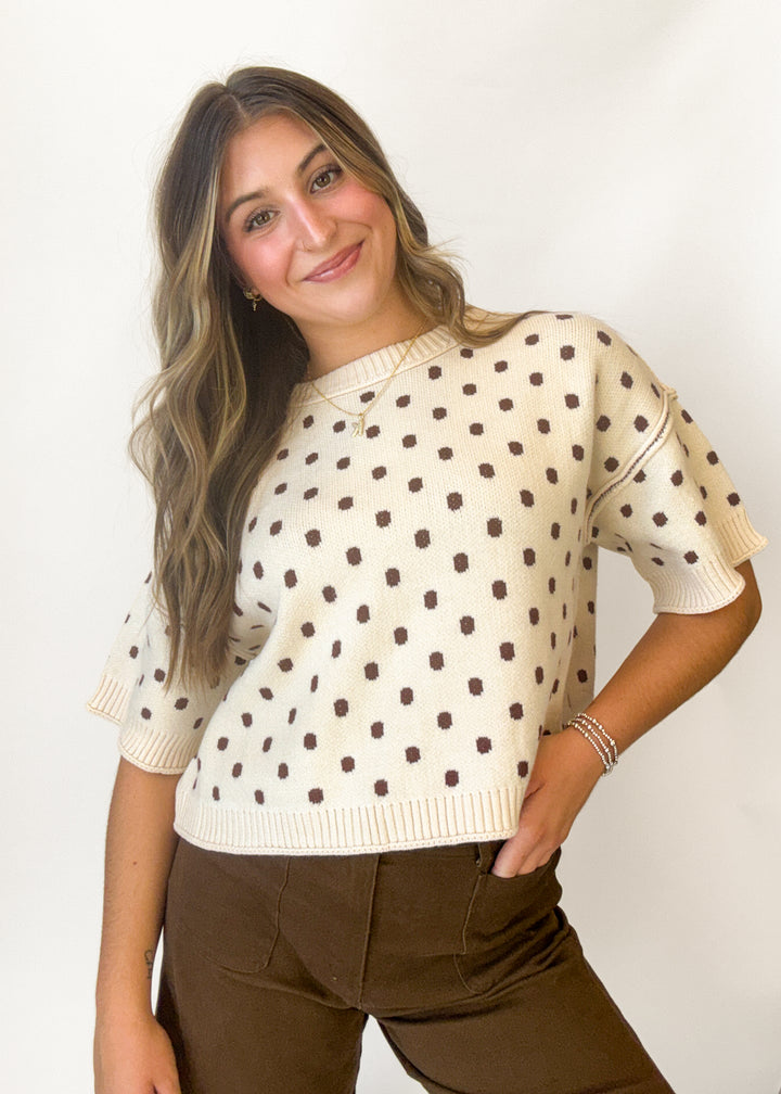 A woman wearing a cream short-sleeve knit sweater with brown polka dots and brown pants, posing against a plain white background.