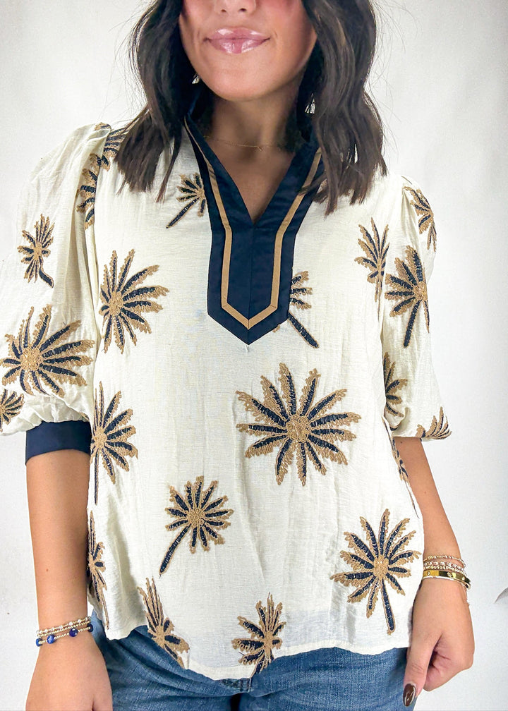 Woman wearing a white blouse with gold floral patterns on a plain background