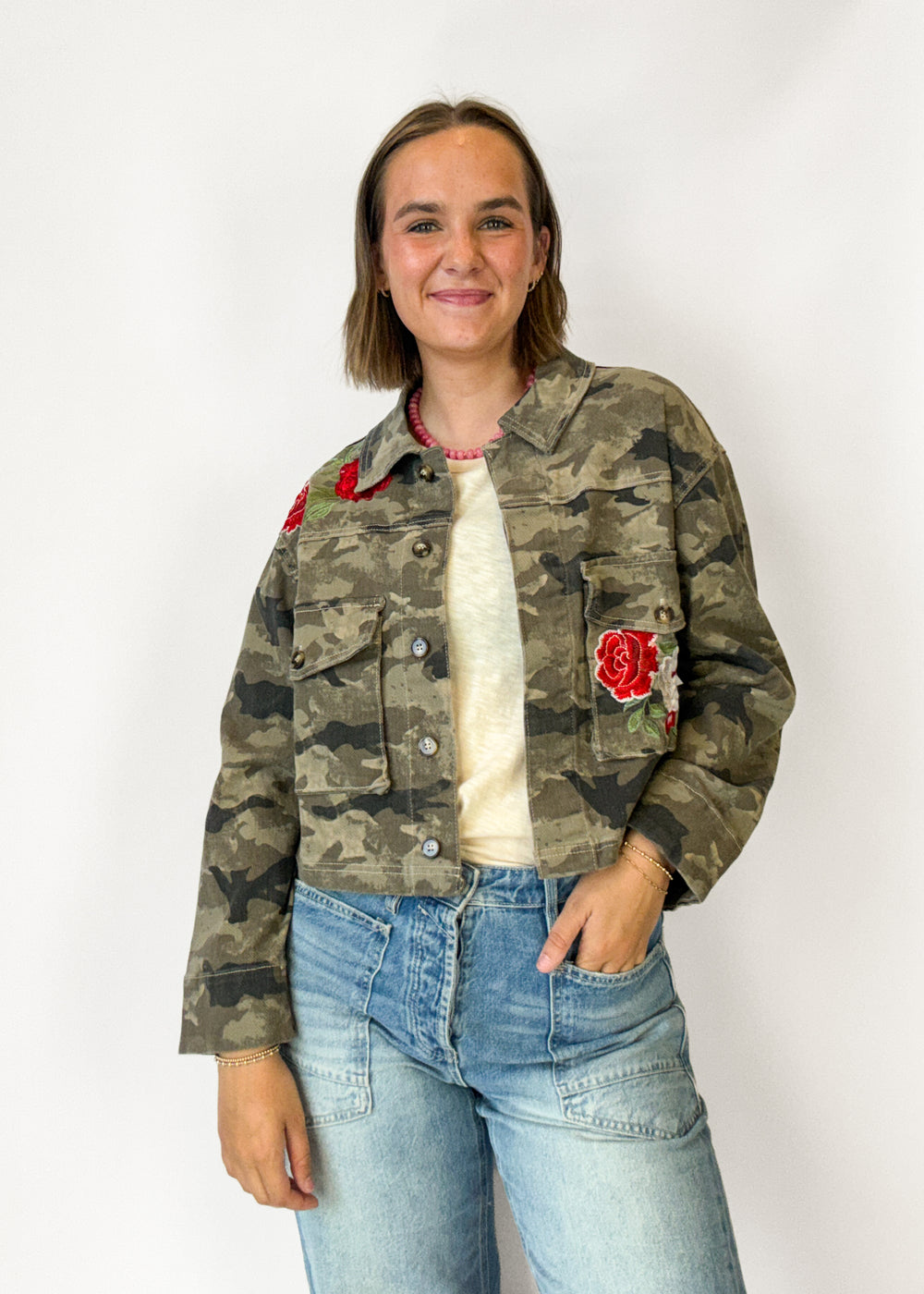 A woman wearing a cropped camouflage denim jacket with red rose patches on the chest, a cream t-shirt, and light blue jeans, standing against a plain white background.