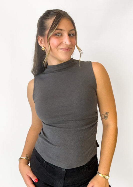 A woman wearing a dark gray sleeveless mock neck top and black pants, standing against a plain white background.