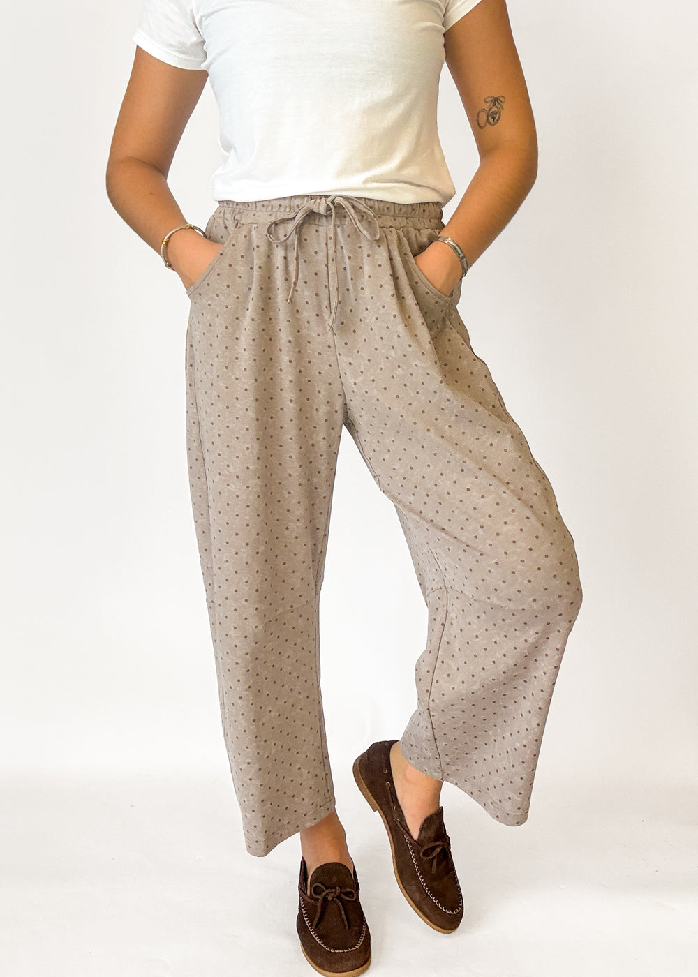 A person wearing loose, wide-leg cropped pants with a small repeating dot pattern in beige and brown, paired with a white top and brown loafers, against a plain light background.