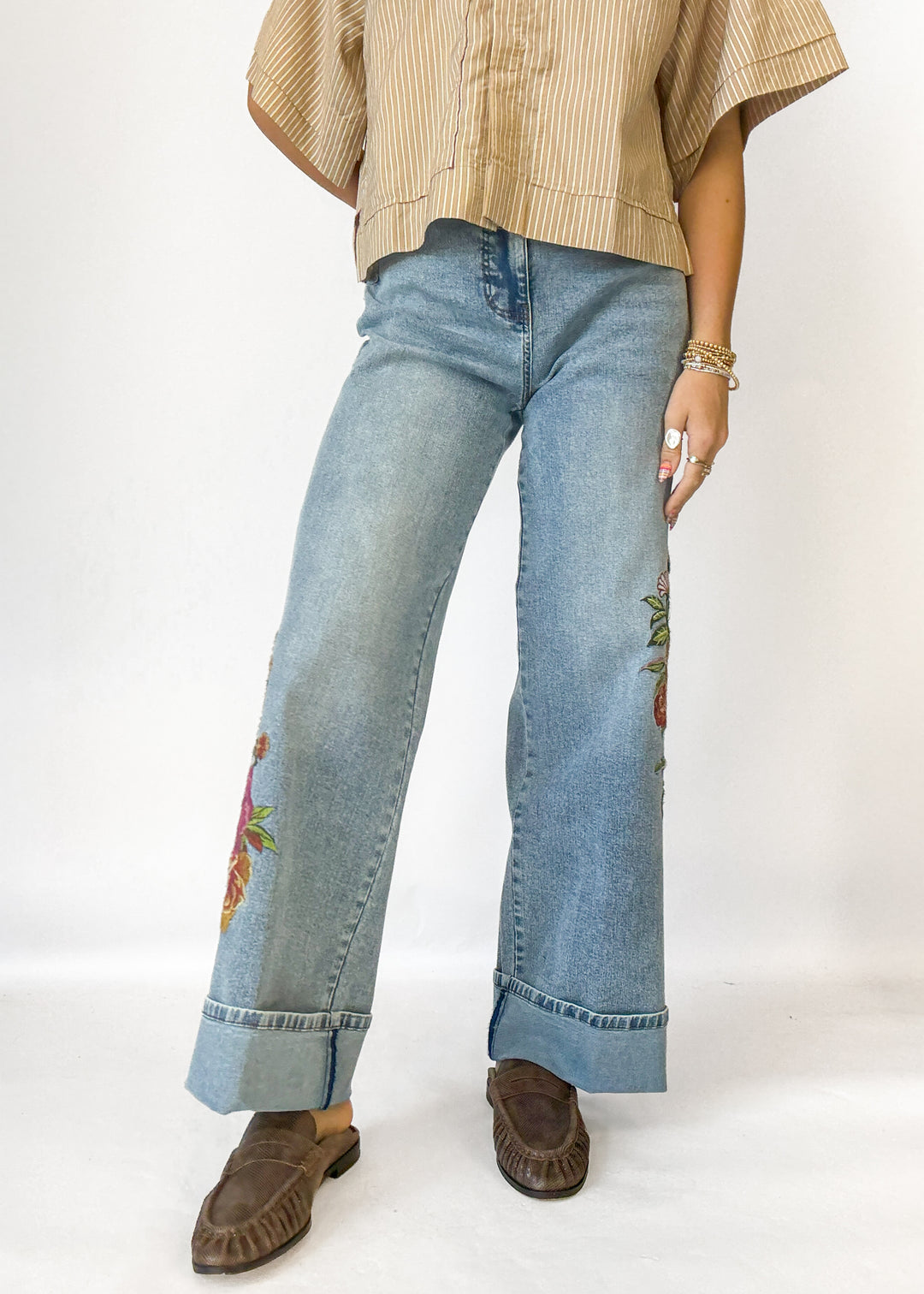 A person wearing light blue wide-leg denim jeans with colorful floral embroidery on the lower legs