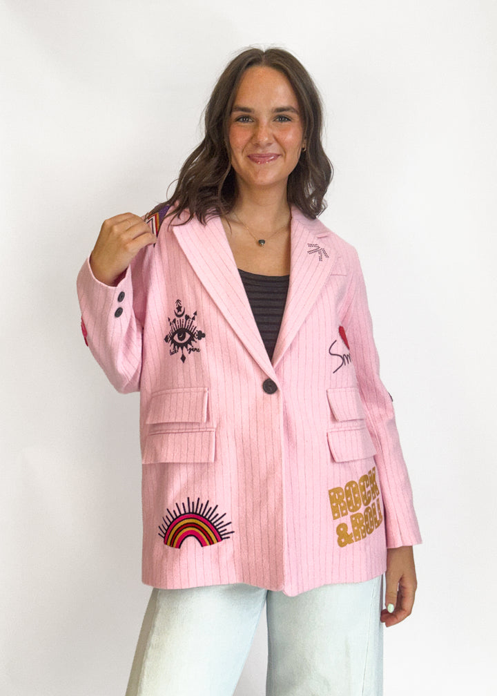A woman modeling a light pink pinstriped blazer with various colorful embroidered patches,