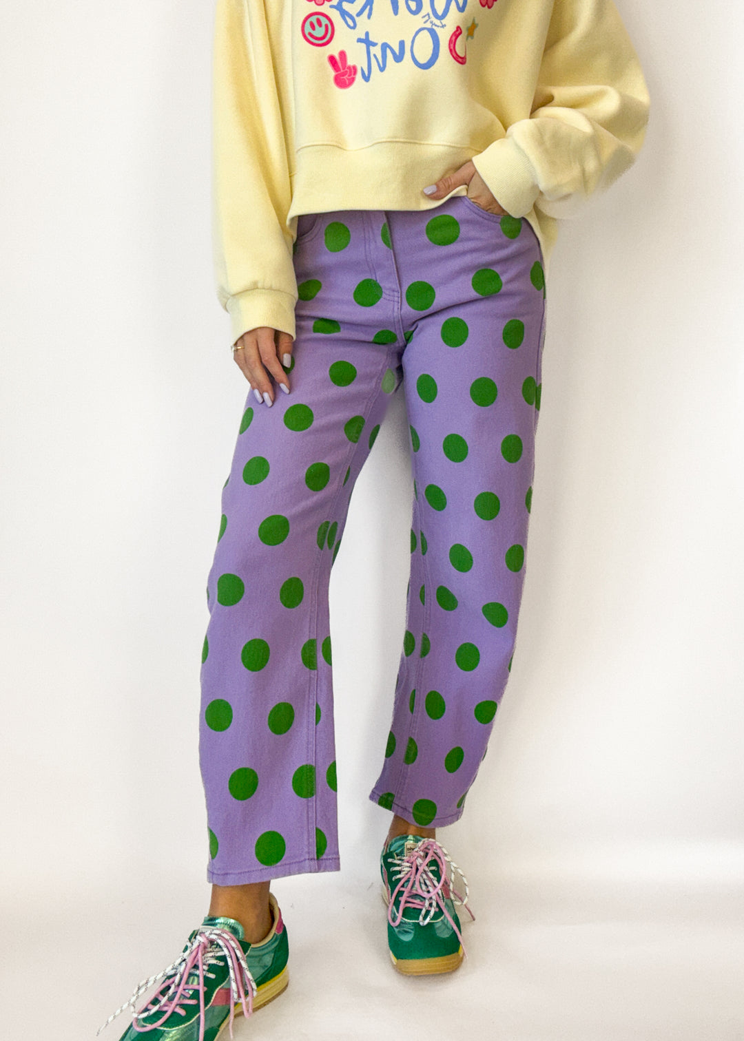 Person wearing a yellow sweatshirt with colorful text, purple pants with green polka dots, and green sneakers on a white background.